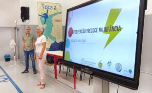 Uma pessoa aparenta estar a falar. Ao lado está um ecrã com uma projeção na qual se lê “Como é que as crianças se desenvolvem?”. A restante informação, no ecrã, é ilegível.