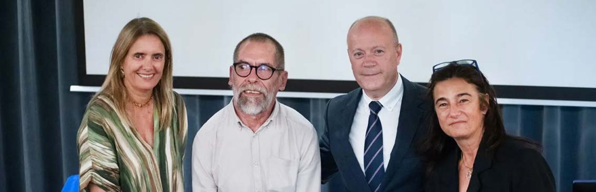Quatro pessoas junto de uma mesa no que aparenta ser um pequeno palco. Uma das pessoas segura uma folha azul que tem em letras brancas a expressão “Diploma de Reconhecimento”. Uma outra pessoa segura um saco azul com a expressão “Voluntariado Porto” em letras brancas.