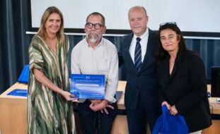 Quatro pessoas junto de uma mesa no que aparenta ser um pequeno palco. Uma das pessoas segura uma folha azul que tem em letras brancas a expressão “Diploma de Reconhecimento”. Uma outra pessoa segura um saco azul com a expressão “Voluntariado Porto” em letras brancas.