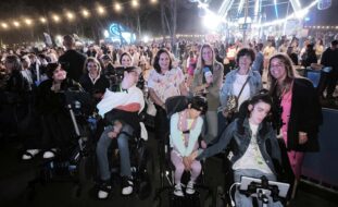 Grupo de uma dezena de pessoas fotografadas em ambiente noturno com várias centenas de pessoas igualmente visíveis na imagem. Ao centro da imagem uma das pessoas segura um microfone identificado com a expressão “Fundação MEO”.