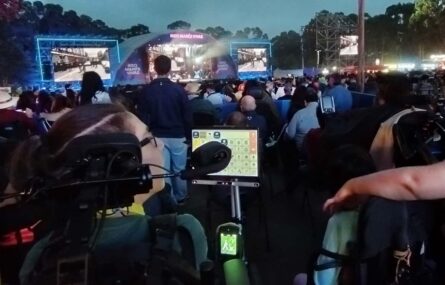 Fotografia noturna de um palco com várias projeções multimédia (no centro e zonas laterais). Em primeiro plano, de costas, uma pessoa sentada em cadeira de rodas usa sistema de comunicação digital.