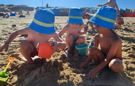 Grupo de crianças na praia, em brincadeira na areia. Ao fundo são visíveis algumas barracas coloridas. Os rostos visíveis das crianças foram digitalmente desfocados.