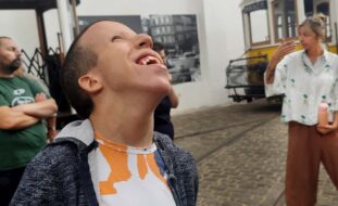 Fotografia de uma pessoa sorridente (aparentemente a olhar para o céu). Surgem ainda outras pessoas na imagem e, ao fundo, é parcialmente visível um dos tradicionais elétricos da cidade do Porto.