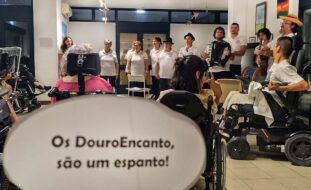 Grupo de pessoas, em espaço interior, aparentemente a cantar. Uma das pessoas segura um acordeão e outra uma viola. EM primeiro plano um pedaço de papel, recortado, com a frase “Os Douro Encanto são um espanto!”