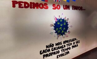 Fotografia de uma parede com uma mensagem (em letras recortadas). A mensagem diz: Pedimos só um favor... Não nos apressem. Cada criança tem o seu próprio tempo para evoluir. Adicionalmente surge uma imagem estilizada do Planeta Terra com várias mãos impressas a verde e diversos desenhos colados.