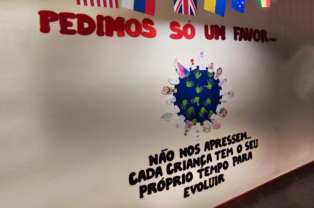 Fotografia de uma parede com uma mensagem (em letras recortadas). A mensagem diz: Pedimos só um favor... Não nos apressem. Cada criança tem o seu próprio tempo para evoluir. Adicionalmente surge uma imagem estilizada do Planeta Terra com várias mãos impressas a verde e diversos desenhos colados.