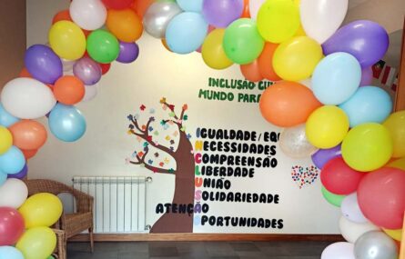 Fotografia de uma parede com uma árvore e várias folhas. Surgem impressas várias palavras e expressões que completam a palavra “Inclusão”. Em primeiro plano um arco com balões coloridos.