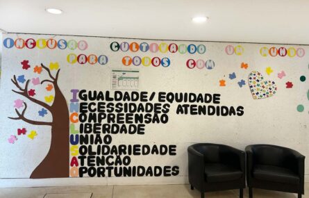 Fotografia de uma parede com uma árvore e várias folhas. Surgem impressas várias palavras e expressões que completam a palavra “Inclusão”. Surgem dois cadeirões na imagem.