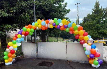 Portão exterior de entrada semi-aberto. Junto do portão está um arco com balões coloridos. Árvores do lado esquerdo e ao fundo da imagem.