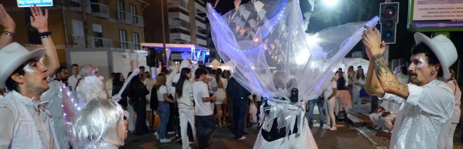 Fotografia noturna de uma rua repleta de pessoas (essencialmente vestidas de branco). Em primeiro plano surgem quatro pessoas em performance artística.