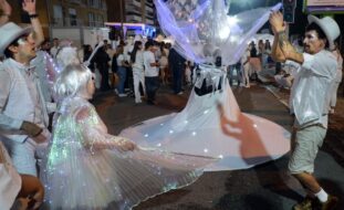 Fotografia noturna de uma rua repleta de pessoas (essencialmente vestidas de branco). Em primeiro plano surgem quatro pessoas em performance artística.
