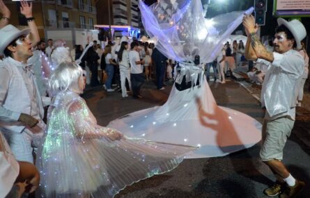 Fotografia noturna de uma rua repleta de pessoas (essencialmente vestidas de branco). Em primeiro plano surgem quatro pessoas em performance artística.