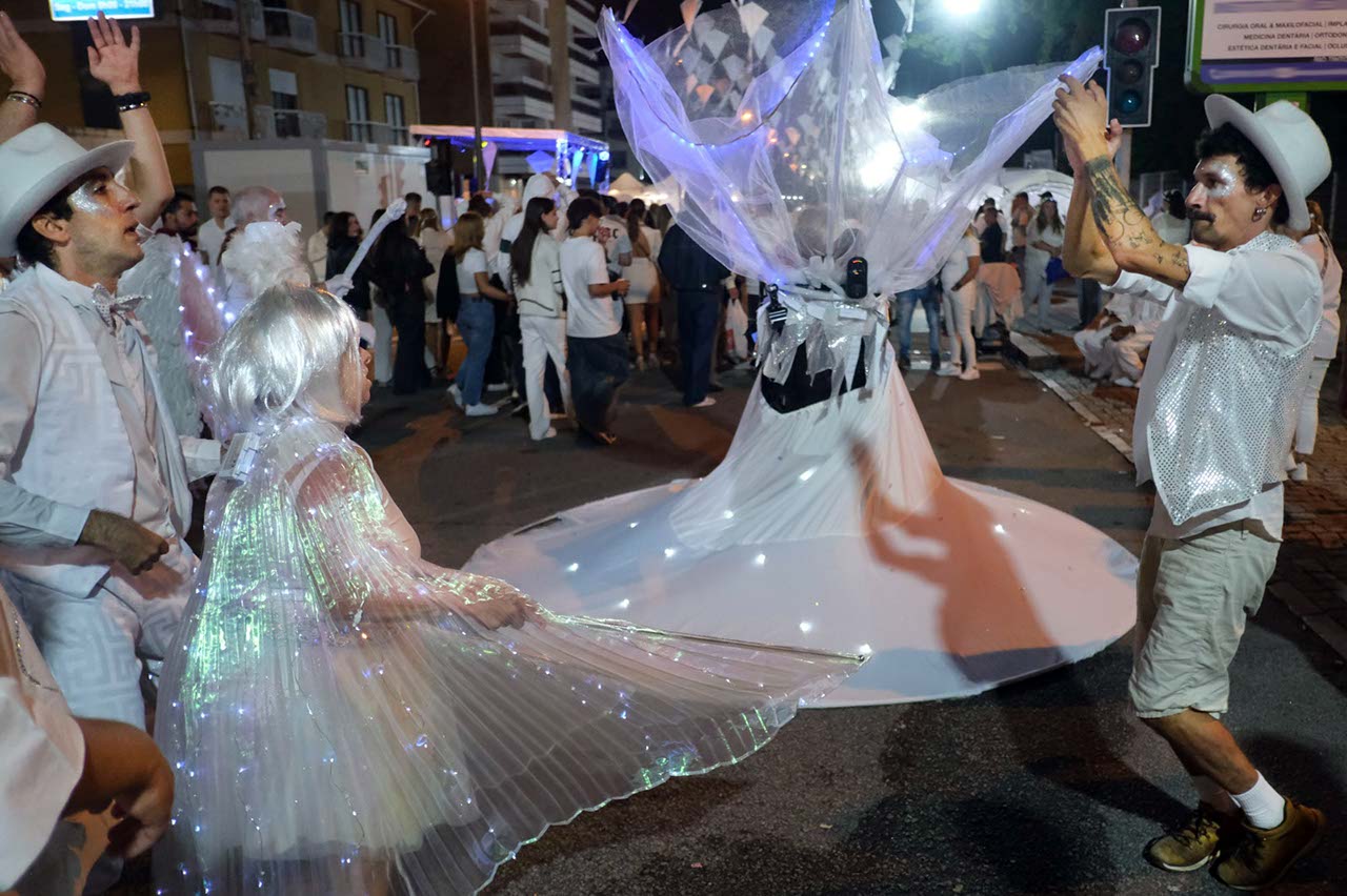 Fotografia noturna de uma rua repleta de pessoas (essencialmente vestidas de branco). Em primeiro plano surgem quatro pessoas em performance artística.
