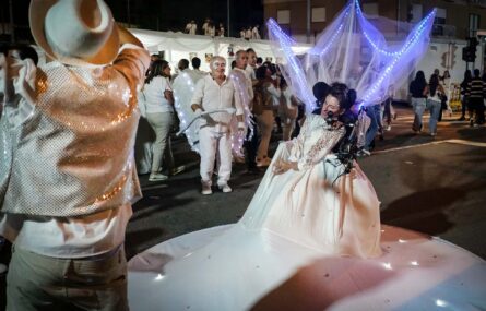 Fotografia noturna de uma rua repleta de pessoas (essencialmente vestidas de branco). Em destaque surgem três pessoas em performance artística.
