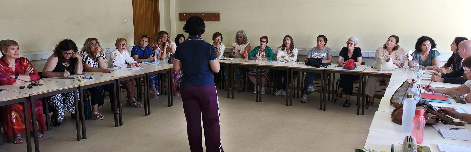 Fotografia de sala interior com cerca de duas dezenas de pessoas sentadas junto de várias mesas (dispostas em formato de U). Ao centro, de pé e de costas, está uma pessoa – que aparenta falar para as restantes. Em cima das mesas estão papéis, malas de mão, carteiras e algumas garrafas de água. Numa das mesas, em destaque, está um pequeno arranjo floral.