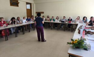 Fotografia de sala interior com cerca de duas dezenas de pessoas sentadas junto de várias mesas (dispostas em formato de U). Ao centro, de pé e de costas, está uma pessoa – que aparenta falar para as restantes. Em cima das mesas estão papéis, malas de mão, carteiras e algumas garrafas de água. Numa das mesas, em destaque, está um pequeno arranjo floral.