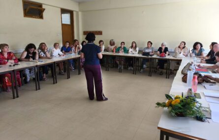Fotografia de sala interior com cerca de duas dezenas de pessoas sentadas junto de várias mesas (dispostas em formato de U). Ao centro, de pé e de costas, está uma pessoa – que aparenta falar para as restantes. Em cima das mesas estão papéis, malas de mão, carteiras e algumas garrafas de água. Numa das mesas, em destaque, está um pequeno arranjo floral.