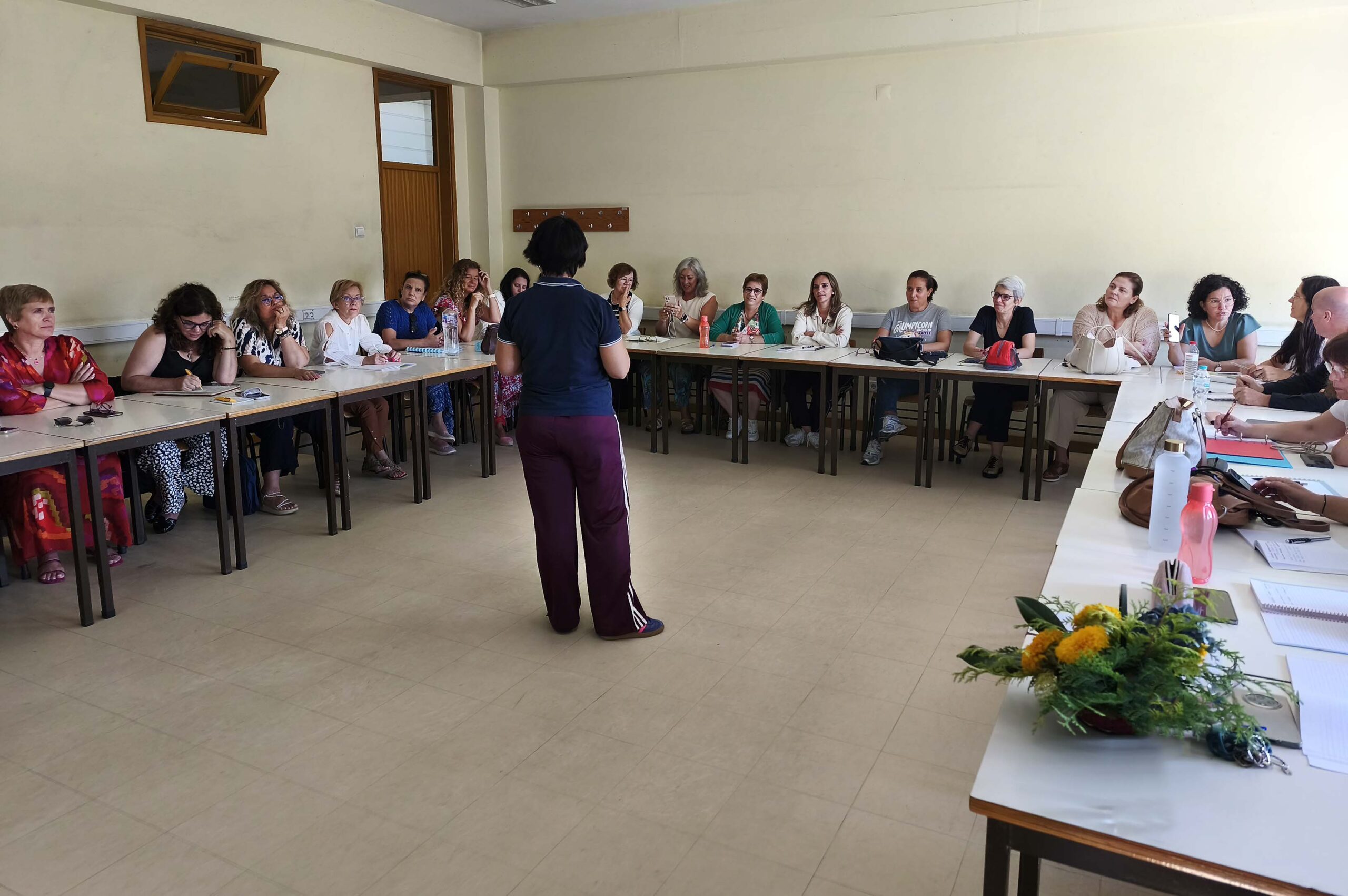 Fotografia de sala interior com cerca de duas dezenas de pessoas sentadas junto de várias mesas (dispostas em formato de U). Ao centro, de pé e de costas, está uma pessoa – que aparenta falar para as restantes. Em cima das mesas estão papéis, malas de mão, carteiras e algumas garrafas de água. Numa das mesas, em destaque, está um pequeno arranjo floral.
